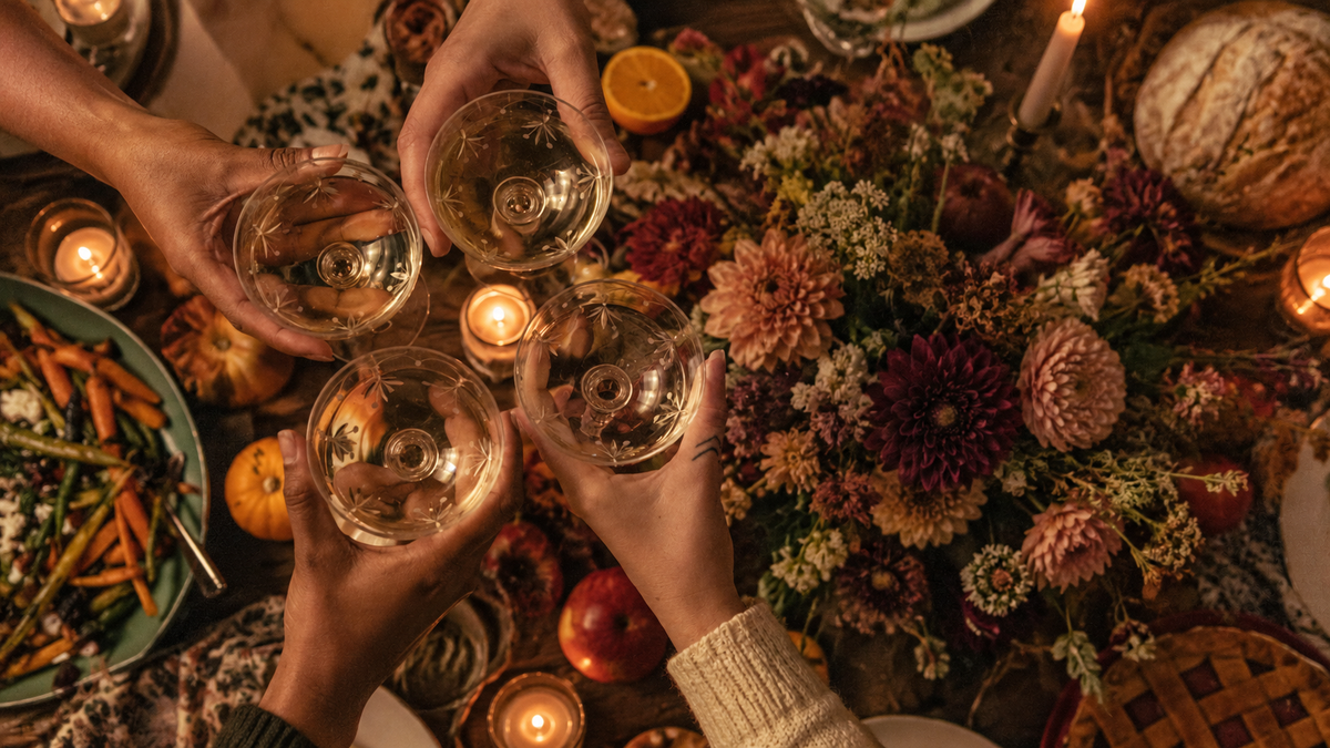 Four people toasting with a summer and fall themed tablescape. Flowers, bread, and plates of food are in the background.