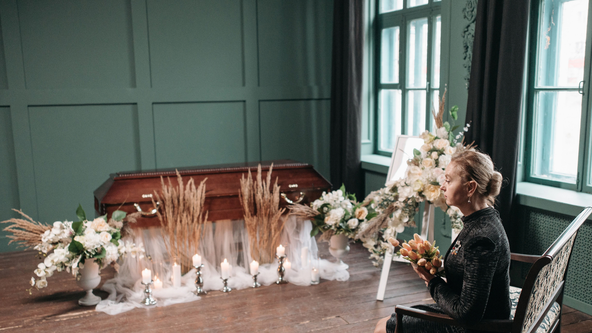 A memorial service or wake with a woman sitting down. The casket is surrounded by flowers and candles.