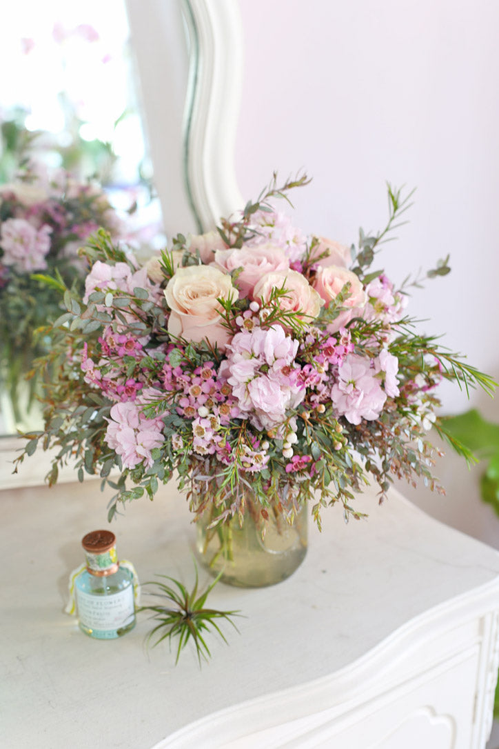 Peach roses, lavender stock, pink wax flowers, and cascading greenery in a glass vase