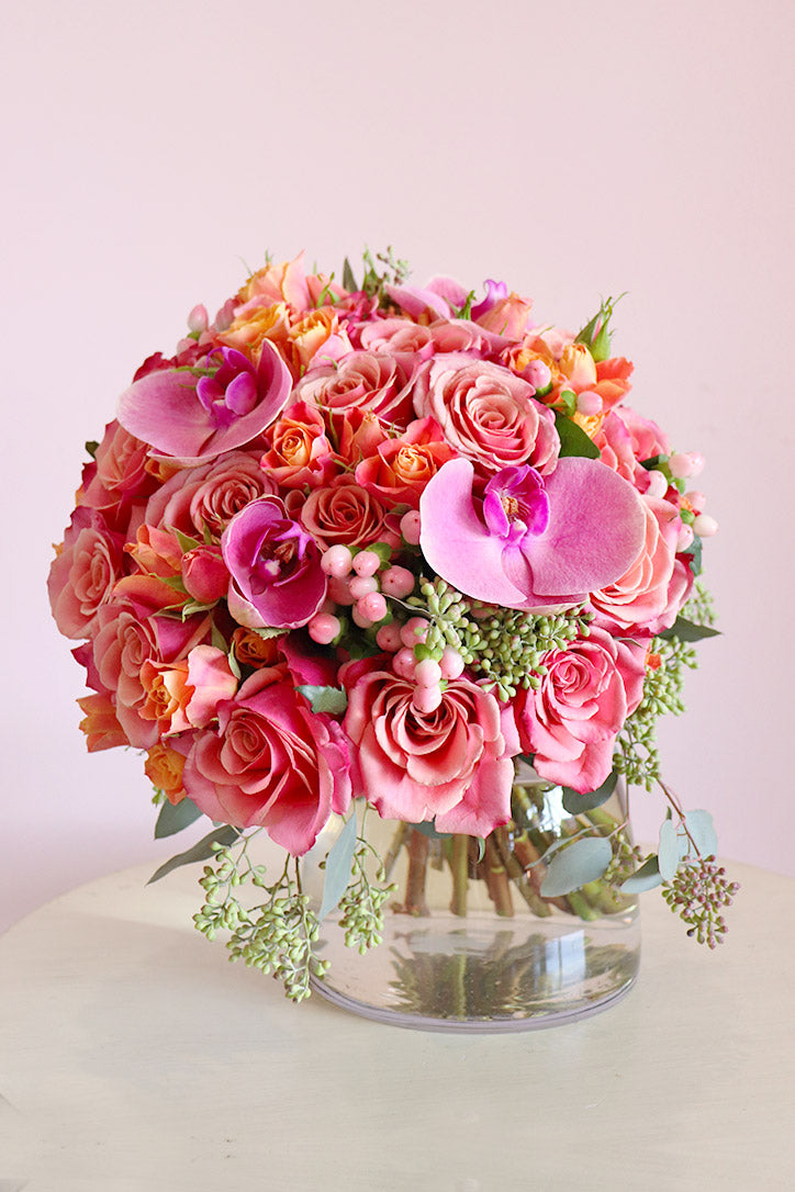 Pink orchids above peach and blush roses with orange spray roses