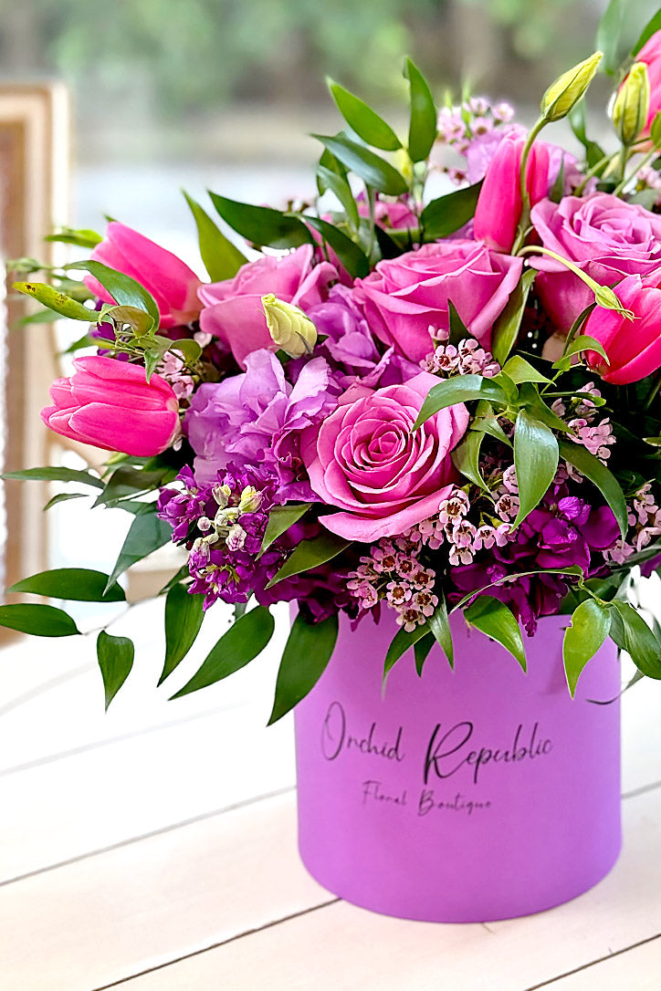 Violet-green hat box filled with mauve, pink and purple flowers and waxflower
