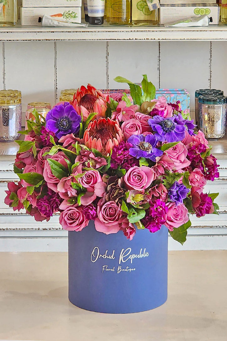 Mauve roses, protea and purple anemones in a navy blue hat box