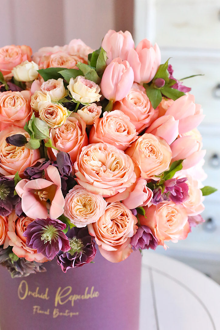 Side view of peach and pink roses, pastel pink tulips, and purple clematis in a plum hatbox