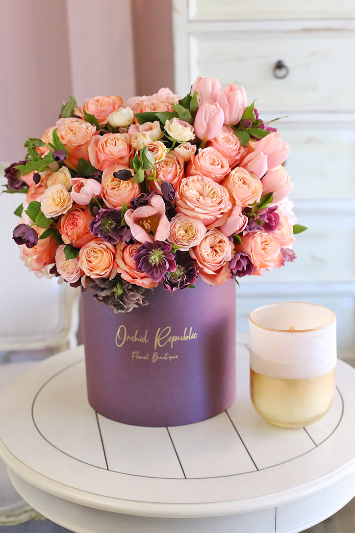 Peach and pink roses, pastel pink tulips, and purple clematis arranged in a plum hatbox
