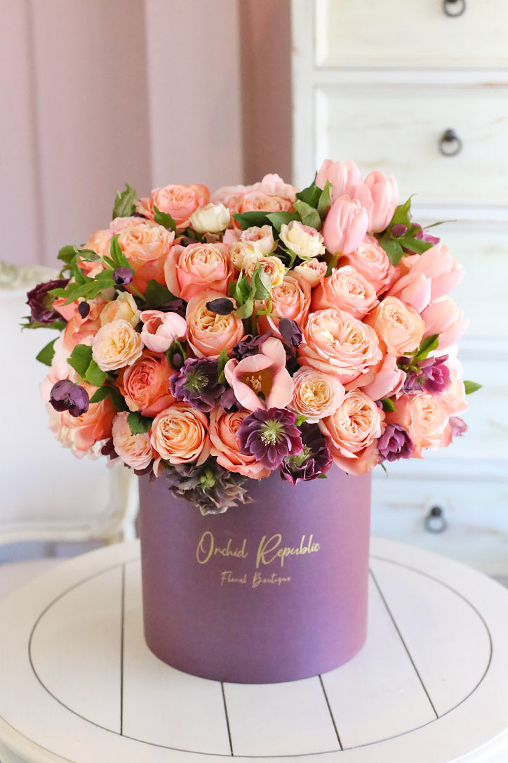 Peach and pink roses, pastel pink tulips, and purple clematis arranged in a plum hatbox
