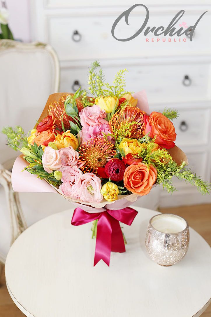 Orange rose bouquet with pincushion protea, yellow tulips, and red ranunculus wrapped in fine paper