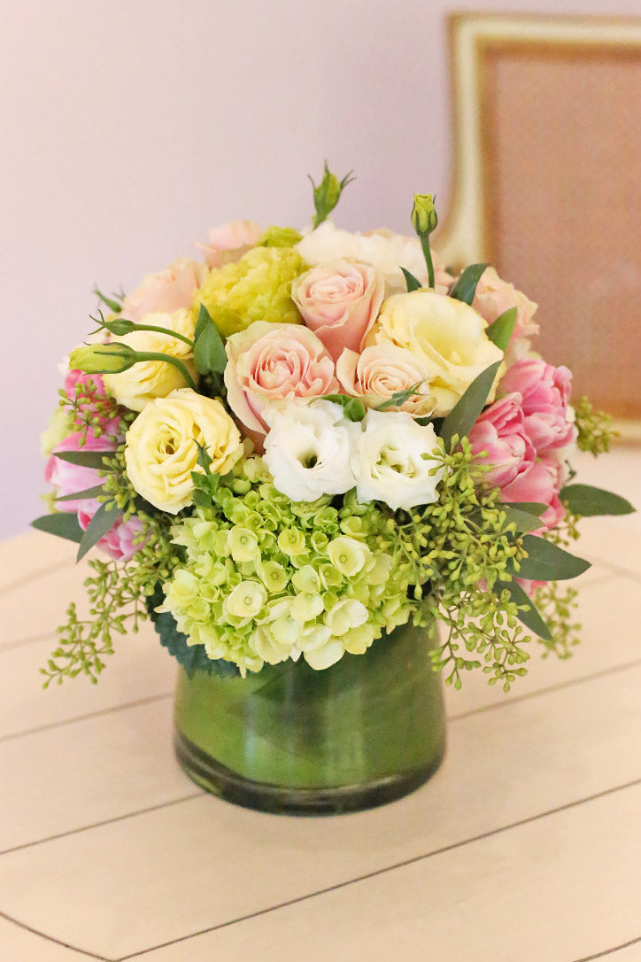 Pink, cream and apple green roses, tulips and hydrangeas in a ti-leaf-lined glass vase