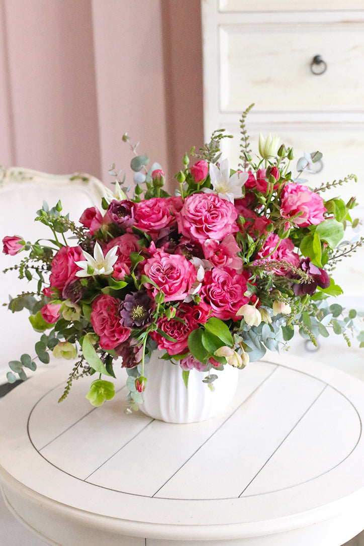 Pink roses, butterfly ranunculus, hellebores, white clematis, and eucalyptus in a white fluted vase
