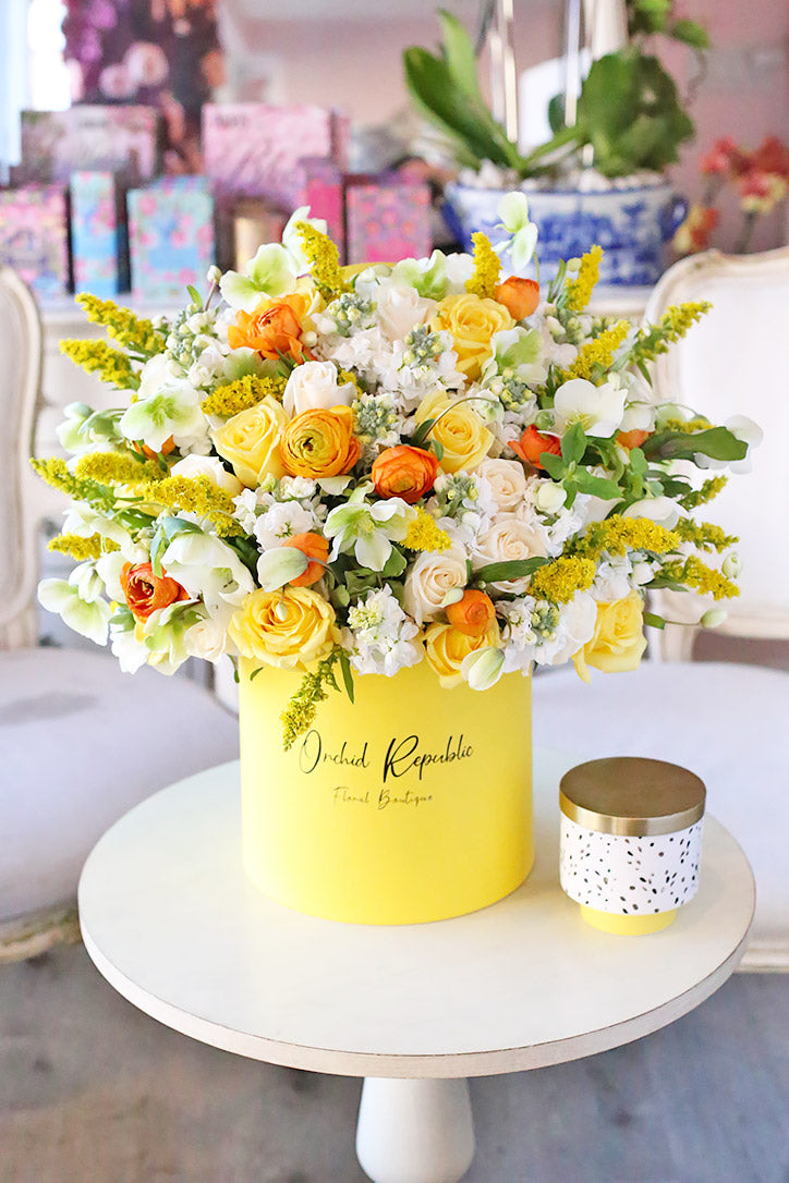 Yellow roses, white stock and orange ranunculus in a yellow hat box