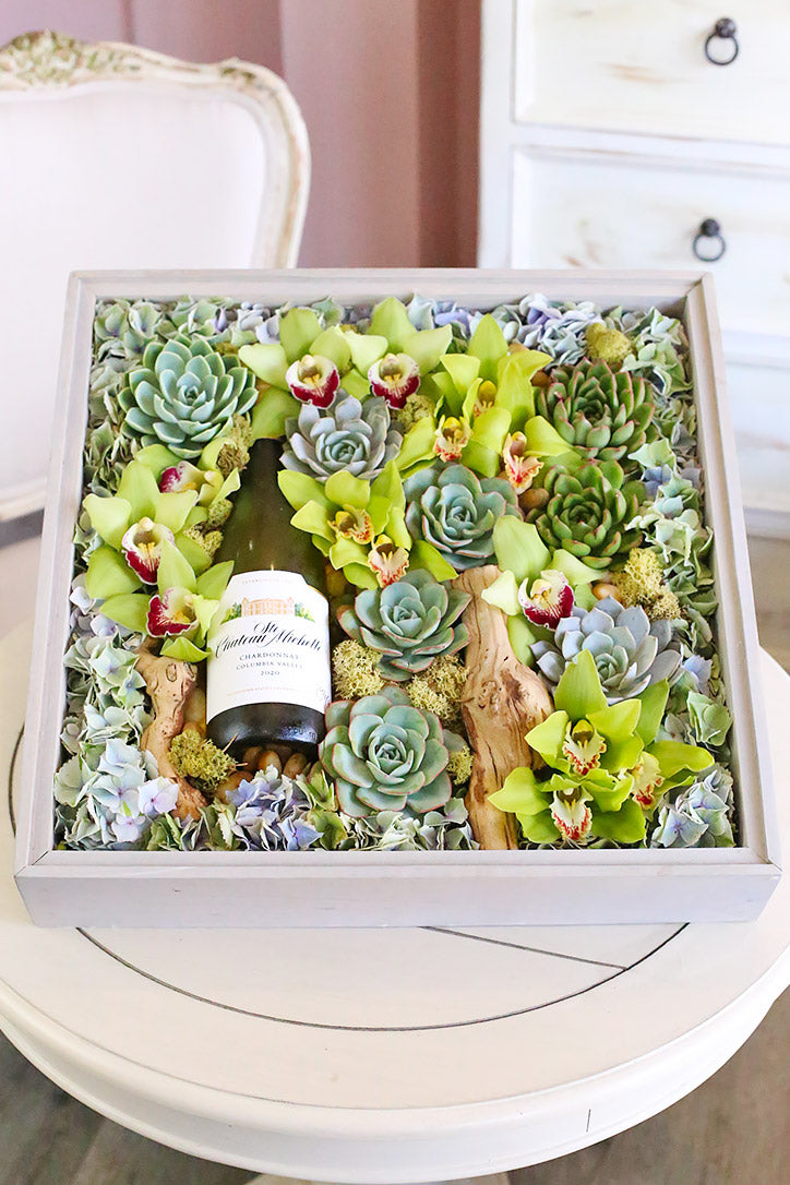 Wooden wine gift box with blue sand, rocks, sculptural wood and a bottle beside fresh greenery.