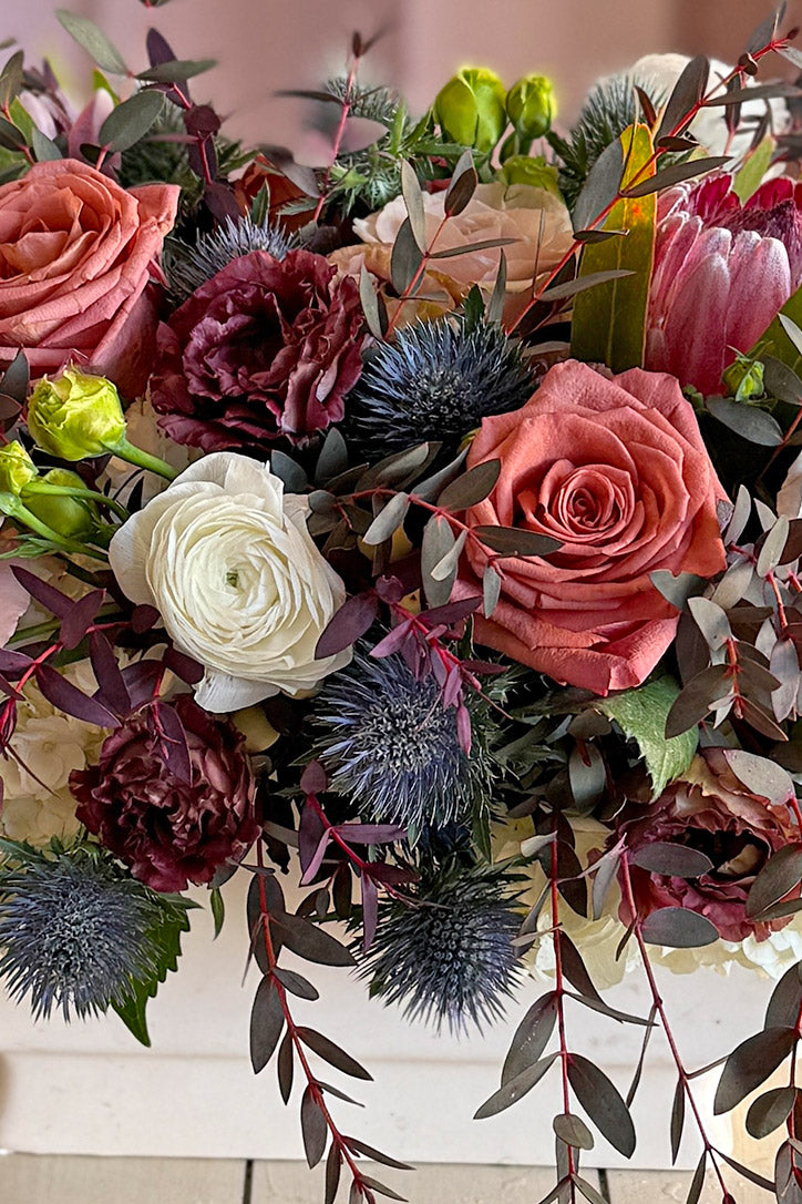 Close-up of neutral roses, white ranunculus, blue thistle and mulberry queen protea